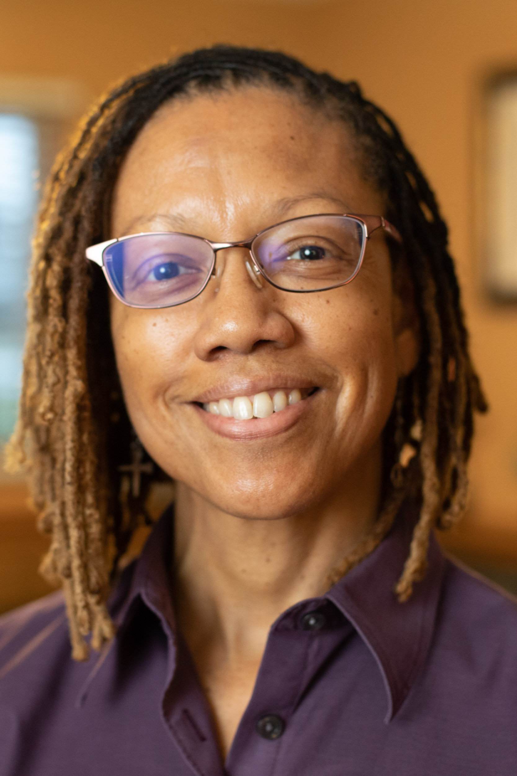 Headshot of a woman with glasses and shoulder-length locs smiling, wearing a dark blouse in a softly lit indoor setting